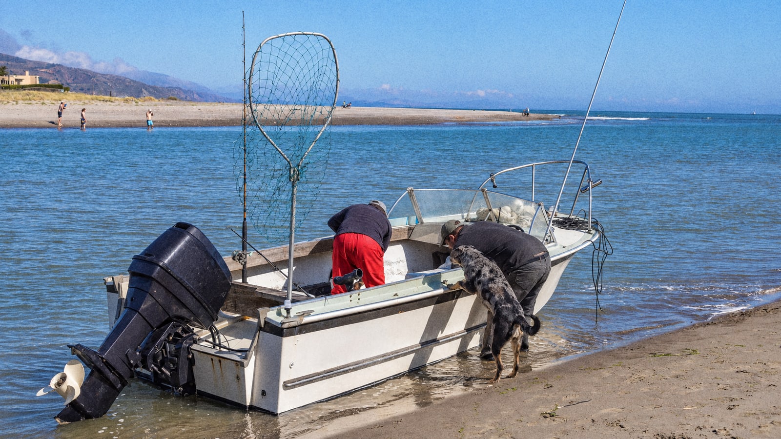 Two men getting a small fishing boat ready on the beach before a morning trip, rods and landing net on board