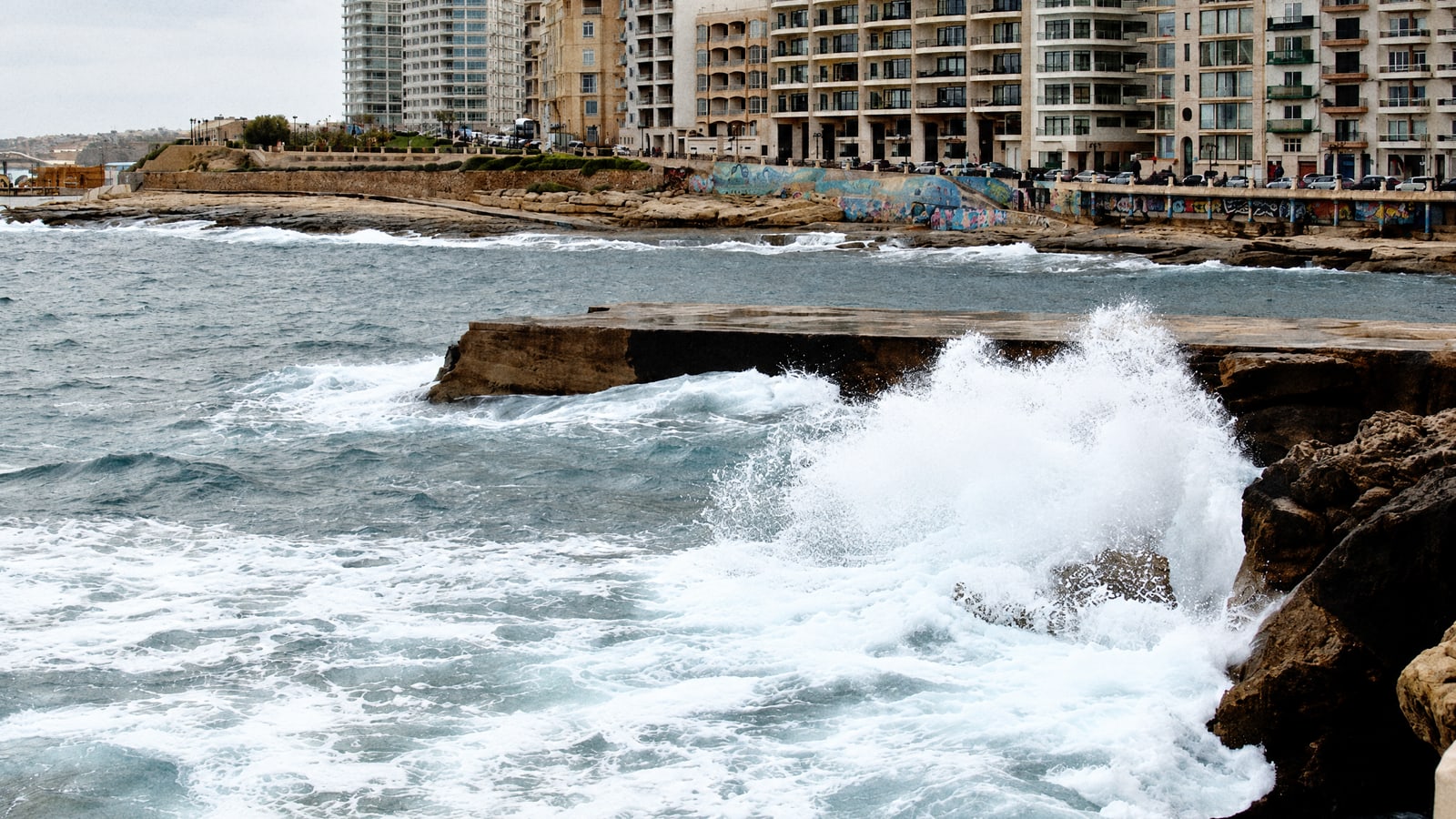 Waves breaking over the rocky shore below the seafront flats of Sliema, Malta, on a windy day