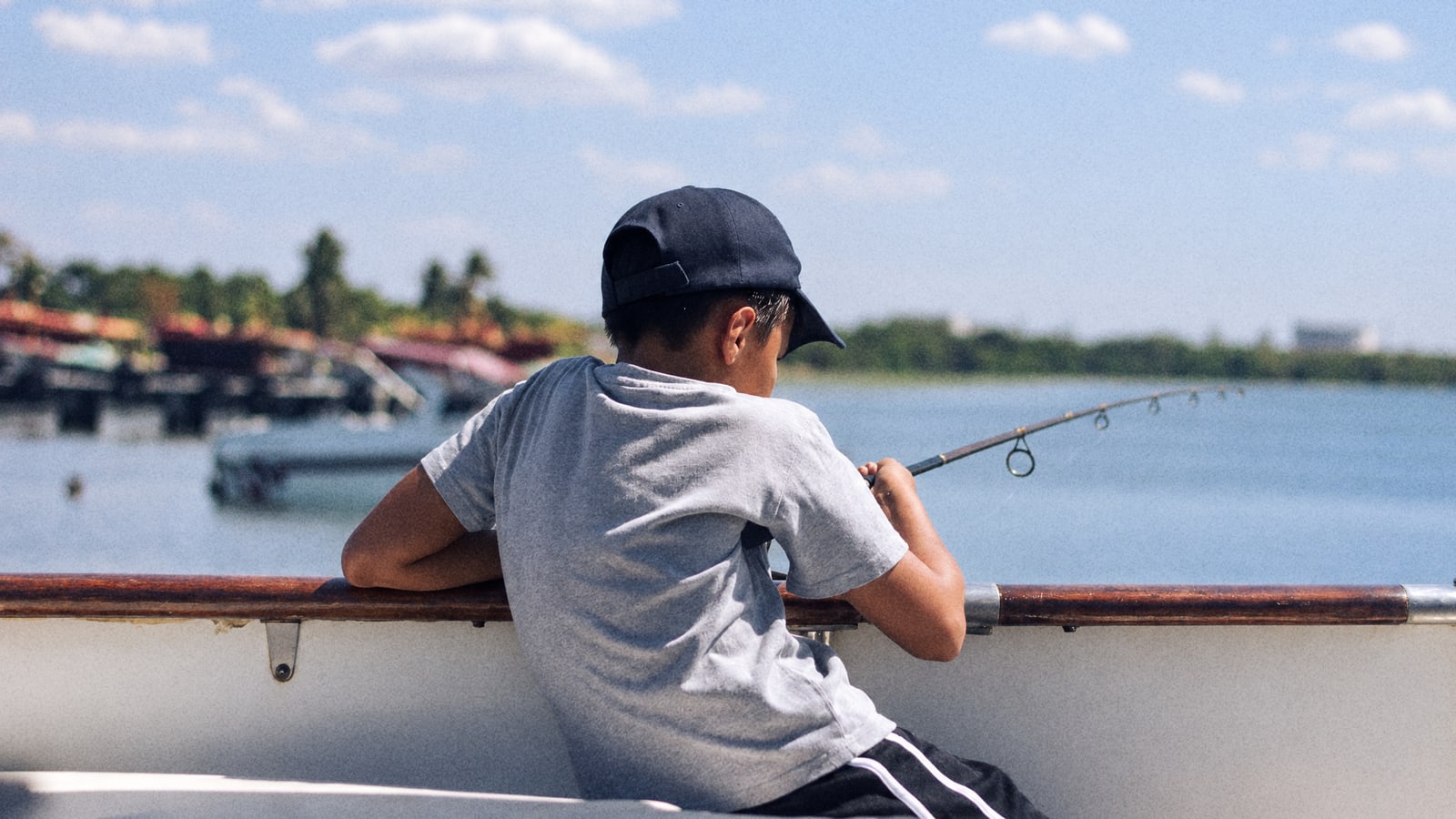 A boy in a cap leaning on the rail of a small boat with a fishing rod over the water